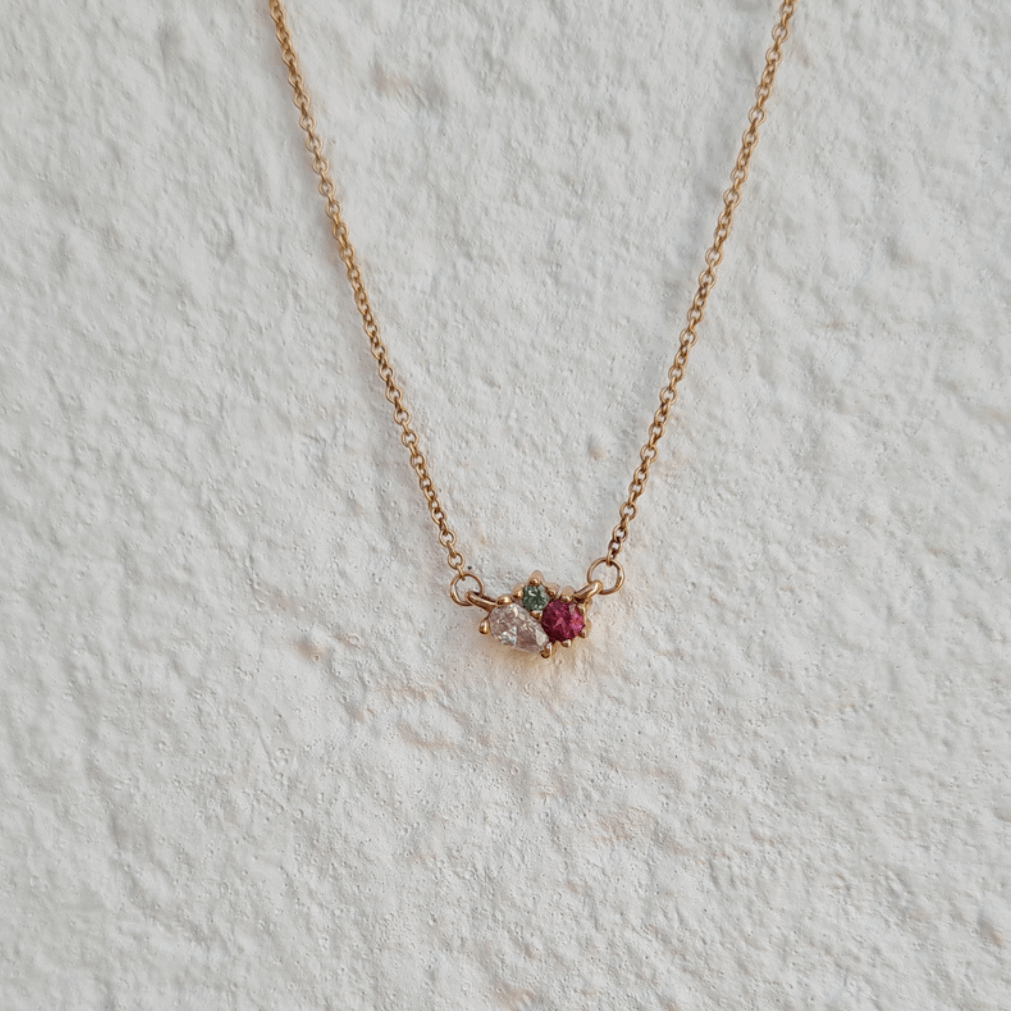 blossom pink and green tourmaline ethically sourced gemstone pendant fine jewellery 18ct recycled rose gold cluster necklace against a white textured wall by designer Emma Hedley
