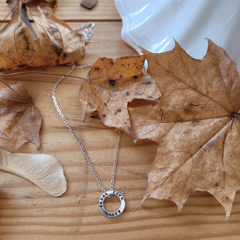 Dainty hoop custom necklace with personalised hand stamped message. Handmade from sterling silver by Emma Hedley Jewellery. Laid on a wooden background surrounded by scattered autumn leaves and sycamore seeds.