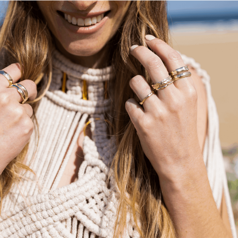 8 Textured recycled yellow gold unique designer stacking rings worn by a woman smiling on a beach with a macrame dress by Emma Hedley Jewellery Claire Collinson Photography