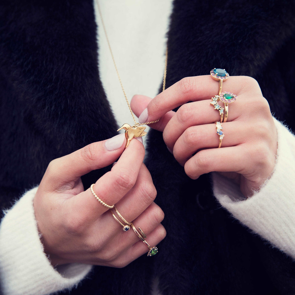 Gold plated silver vermeil hummingbird pendant held in a woman's hands wearing stacks of gold and colourful gemstone rings - By British Fine jewellery designer Emma Hedley