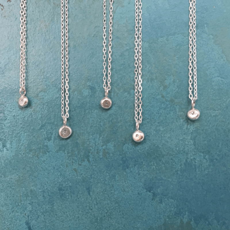 melted initial nugget pendant tiny alphabet charm handmade necklaces hanging on trace chains against a teal background. Handmade in Sterling silver by Emma Hedley Jewellery Designer