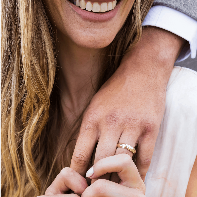 A woman smiling whilst holding a man's hand- wearing a unique Organic Textured 5mm yellow gold wedding ring- by fine jeweller Emma Hedley- Photography by Claire Collinson