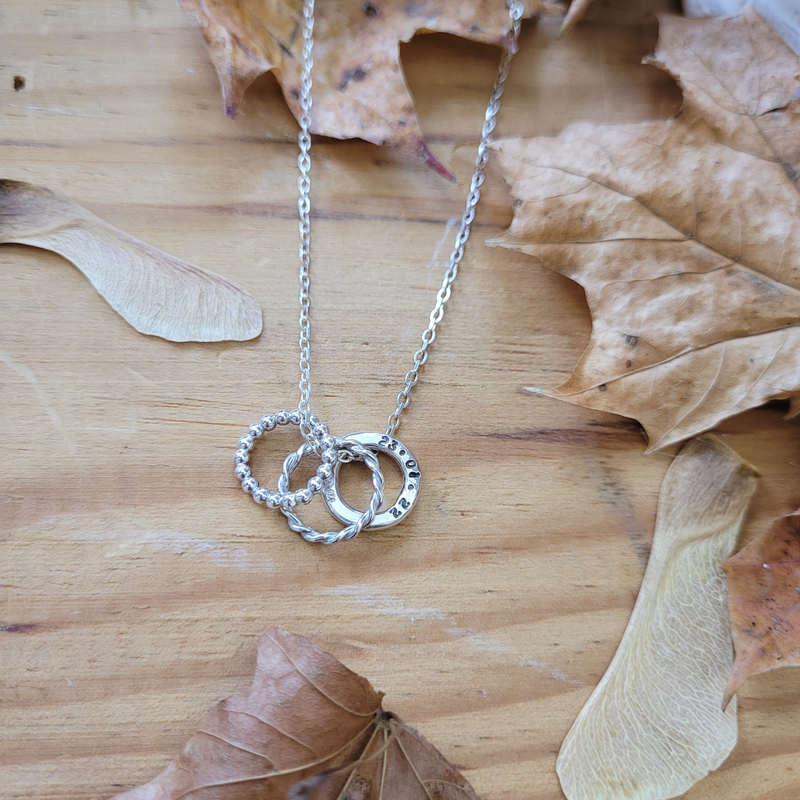 Textured Hoop Necklace with three hoops; one handstamped personalised hoop. Hanging on an 18" trace chain, laid on a wooden background with leaves and sycamore seed scattered around. Handmade by Emma Hedley Jewellery
